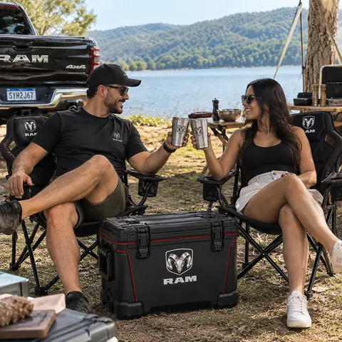 Casal em cadeiras RAM à beira do lago brindando com copos térmicos, cooler RAM na frente